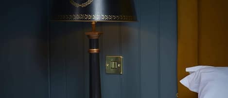 Una mesita de noche de madera con lámpara negra que presenta detalles dorados y un pie decorativo, ubicada junto a una cama con ropa blanca.