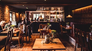 Un restaurant chaleureux avec des tables et des chaises en bois, un bar à l’arrière-plan et un mur de pierre sur l’un des côtés.