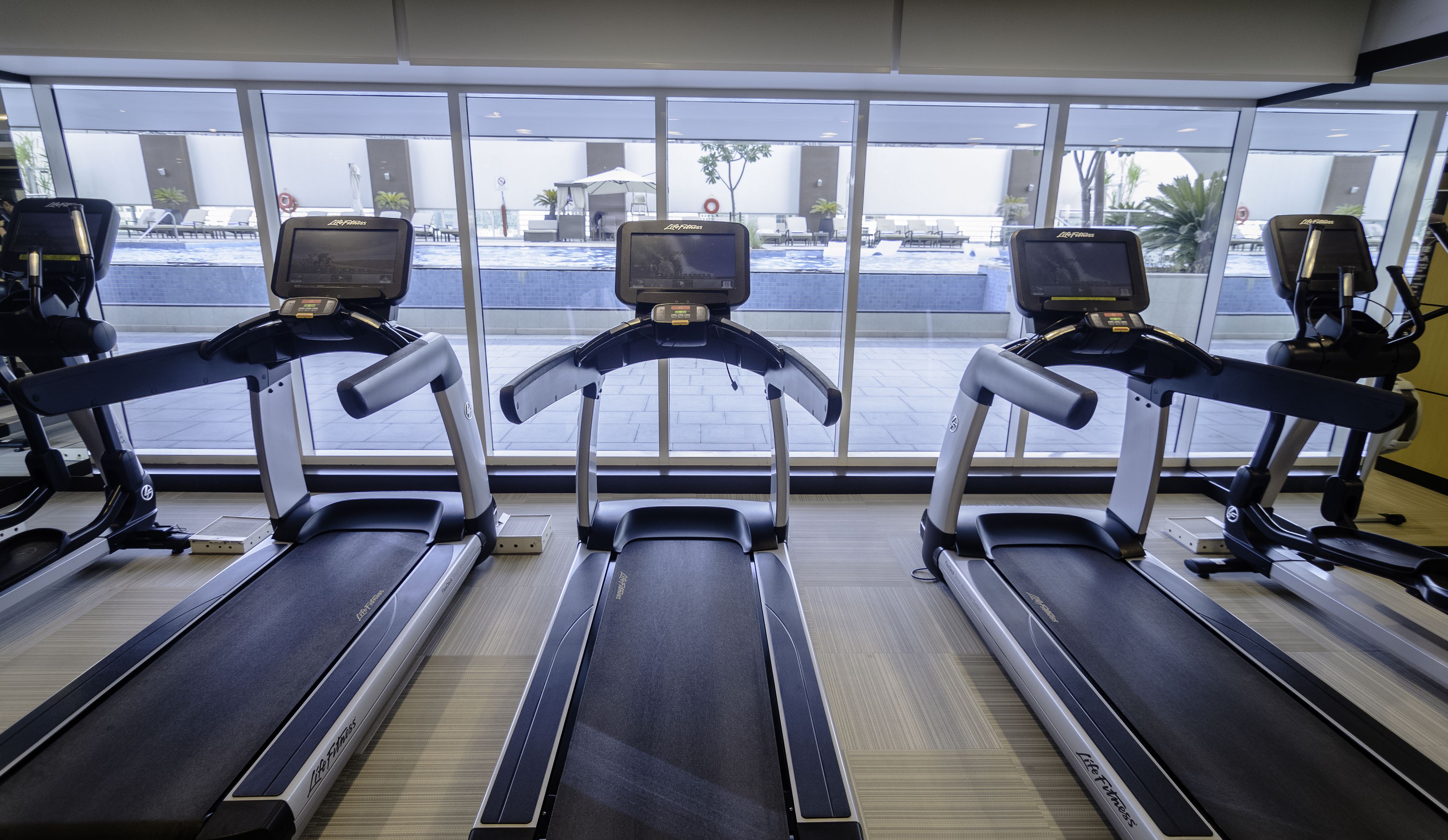 Tres cintas de correr en un gimnasio con ventanas amplias que dan a una zona de piscina.
