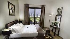Quarto de hotel com uma cama, mesa de cabeceira com abajur, um vaso de flores sobre uma mesinha e um quadro na parede, além de vista para a cidade através de uma grande janela.
