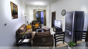 Uma sala de estar moderna com sofá, mesa de centro e televisão. Há também uma geladeira e um espaço de refeições com cadeiras e mesa.