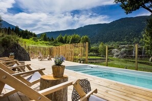 Una terraza de madera con sillones de descanso y piscina, rodeada por una valla de madera y un paisaje montañoso.