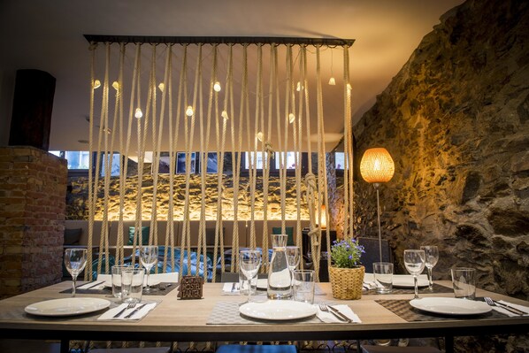 A dining table set with plates, glasses, and cutlery, with a rope curtain overhead and a stone wall in the background. - Mountain Hostel Tarter (El Tarter)