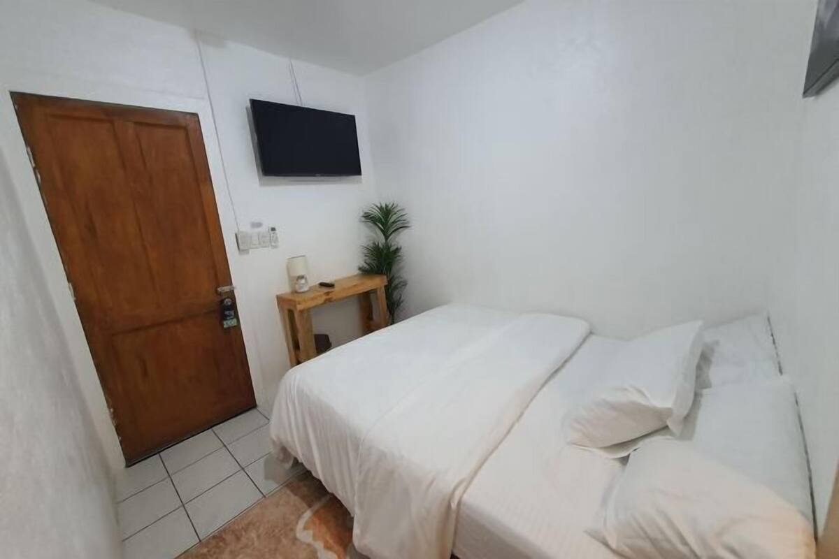 A bedroom with a bed, a wooden door, a small table with a plant, and a TV mounted on the wall.