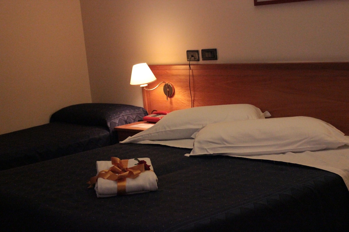 A hotel room with two beds, a nightstand, a lamp, and a wall-mounted clock.