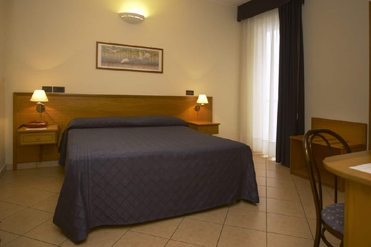 A hotel room with a large bed, two bedside lamps, a wooden headboard, a desk with a chair, and a painting on the wall.