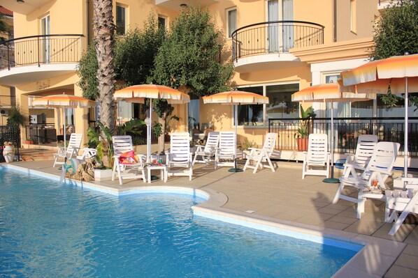 A poolside area with sun loungers and umbrellas, a palm tree, and a building with balconies.