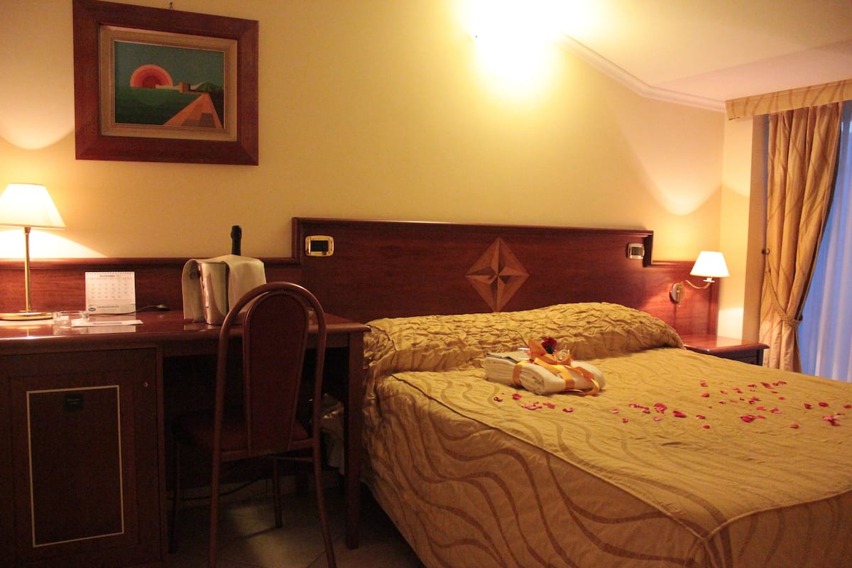 A hotel room with a bed, a desk, a chair, and a nightstand with a lamp.
