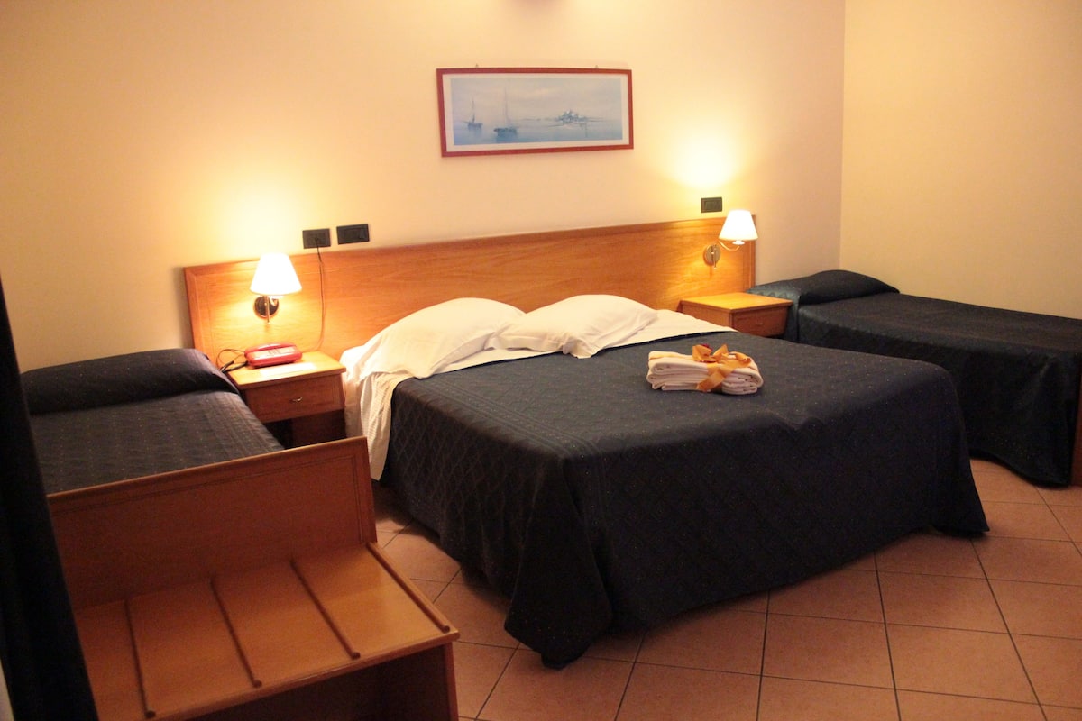 A hotel room with two single beds, a wooden bench, a nightstand, and a wall-mounted lamp.