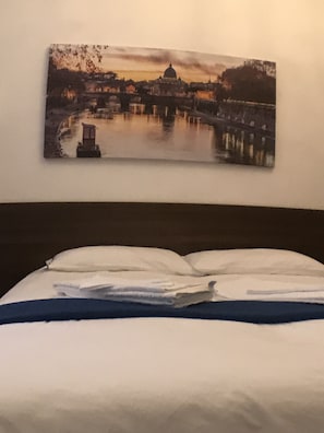 A bed with white bedding and a blue accent, a painting of a cityscape with a river and a bridge, and a plain wall.