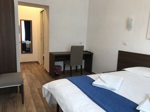 Habitación de hotel con cama, escritorio y silla. Hay un espejo en la pared y una puerta que da a otra habitación.