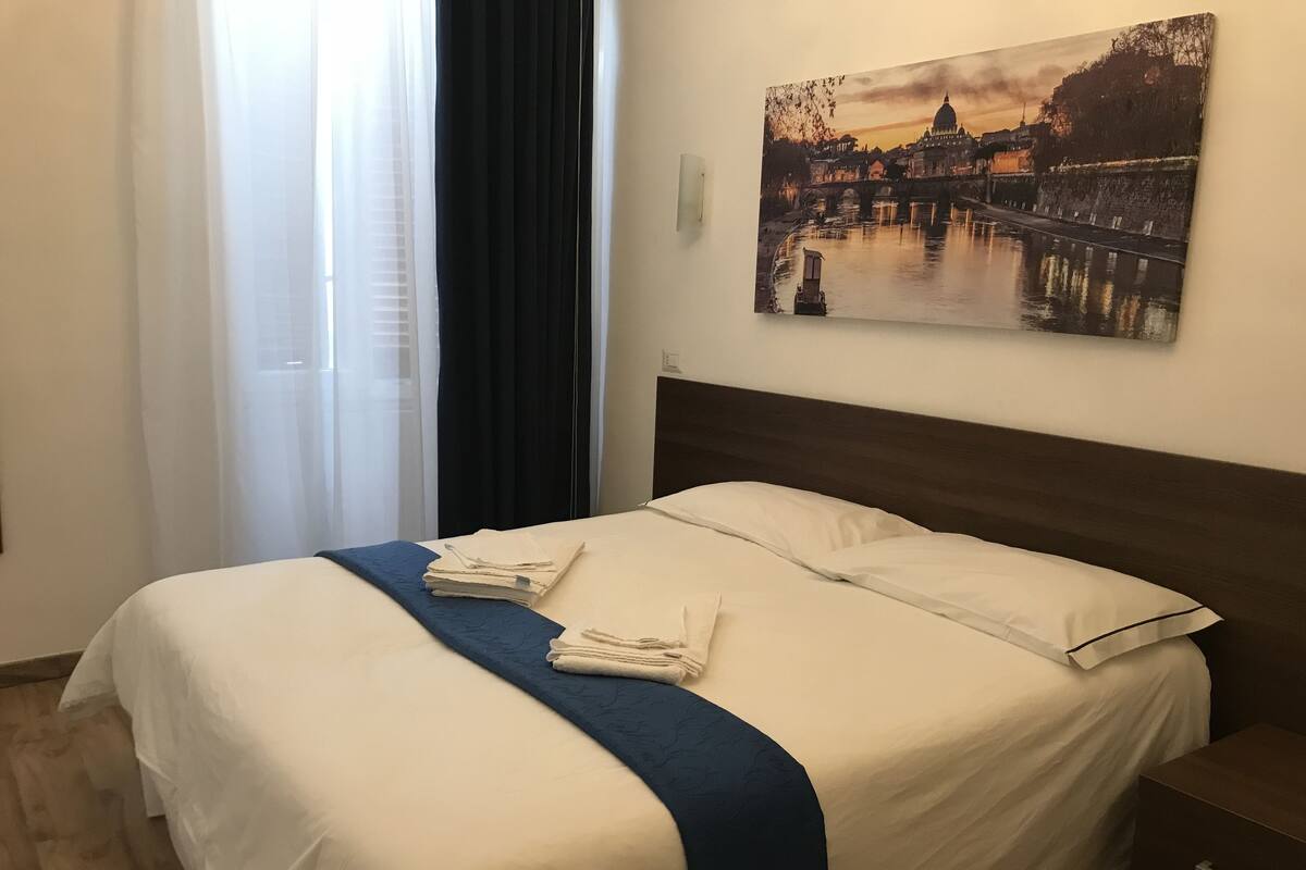 A neatly made bed with white and blue linens, a folded towel, and a painting of a cityscape on the wall.