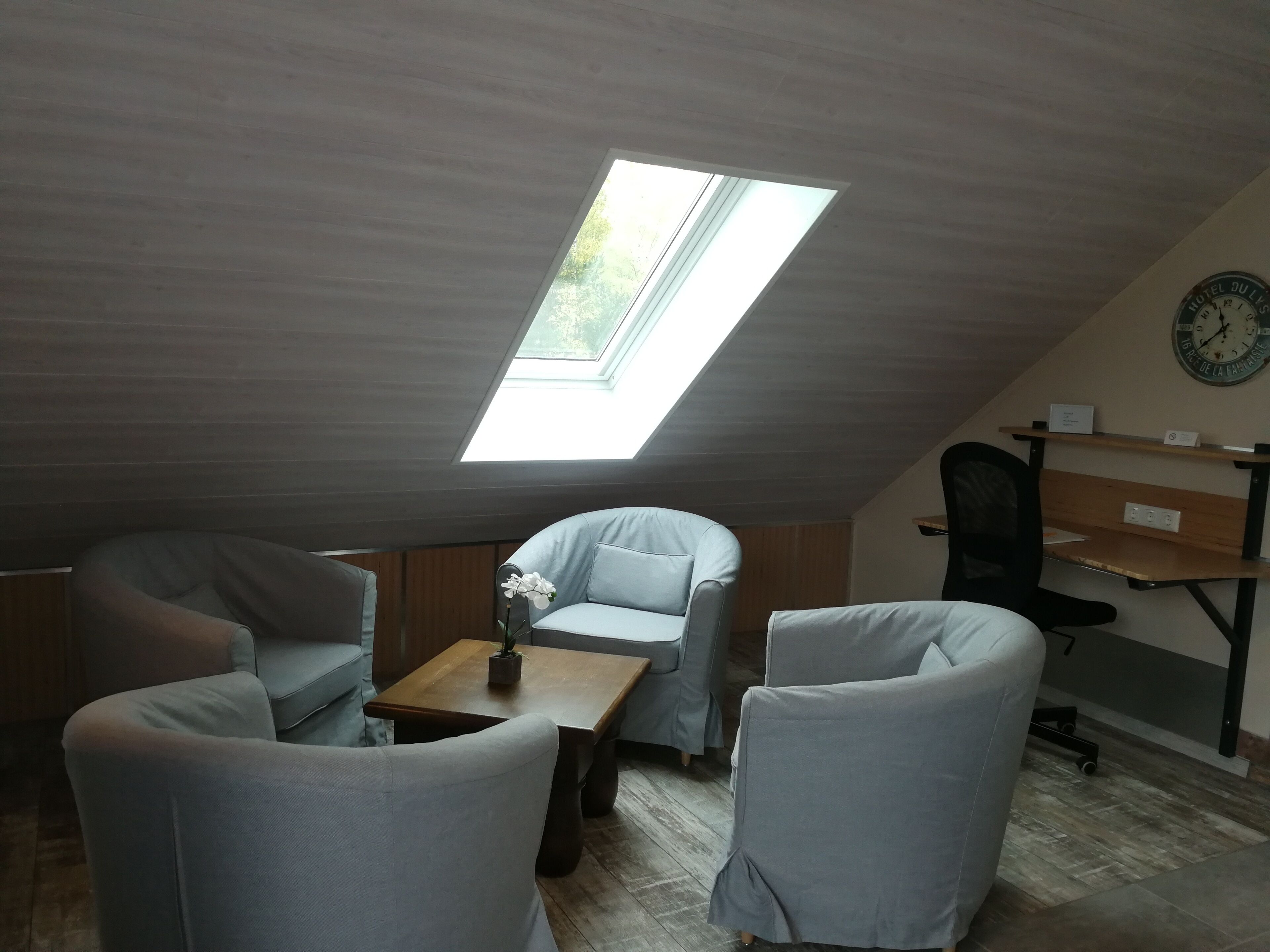 Une pièce mansardée accueillante, dotée d’une lucarne, de deux fauteuils gris, d’une table en bois et d’un bureau avec une horloge.