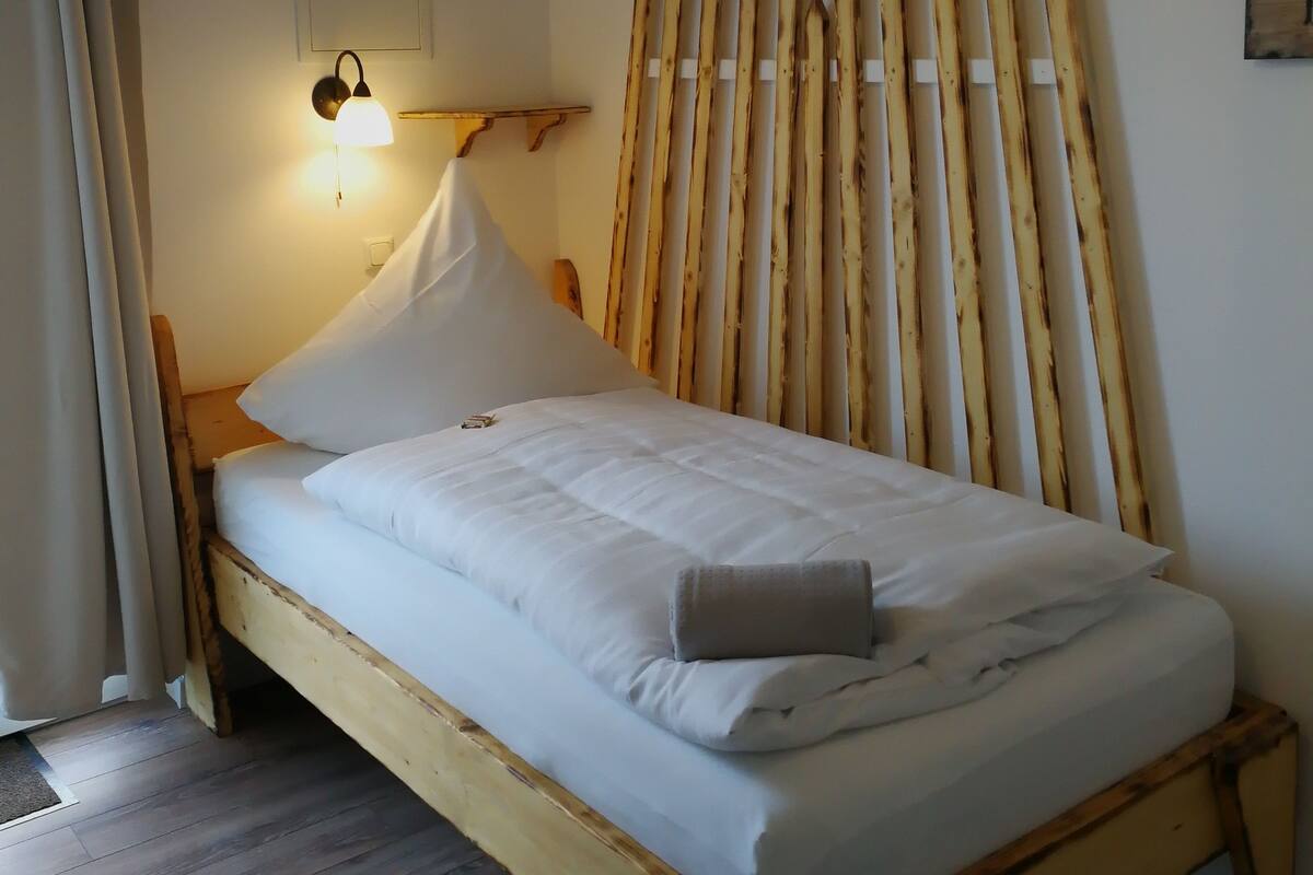 A single bed with white bedding, a wooden headboard, a wall-mounted light, and a wooden slat partition.