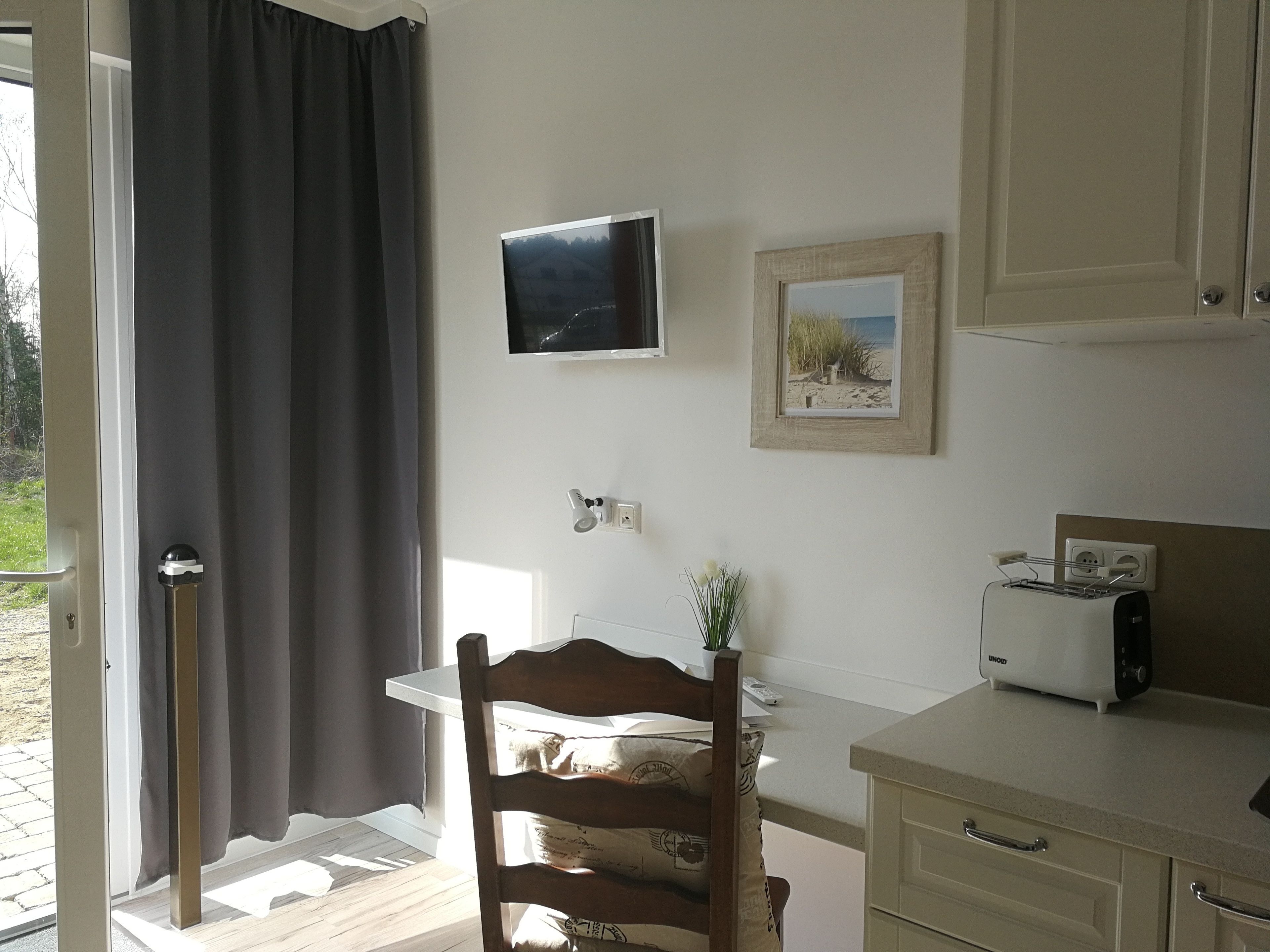 A small kitchen with a table, chair, and a toaster. There is a window with a curtain, a wall-mounted TV, and a framed picture.