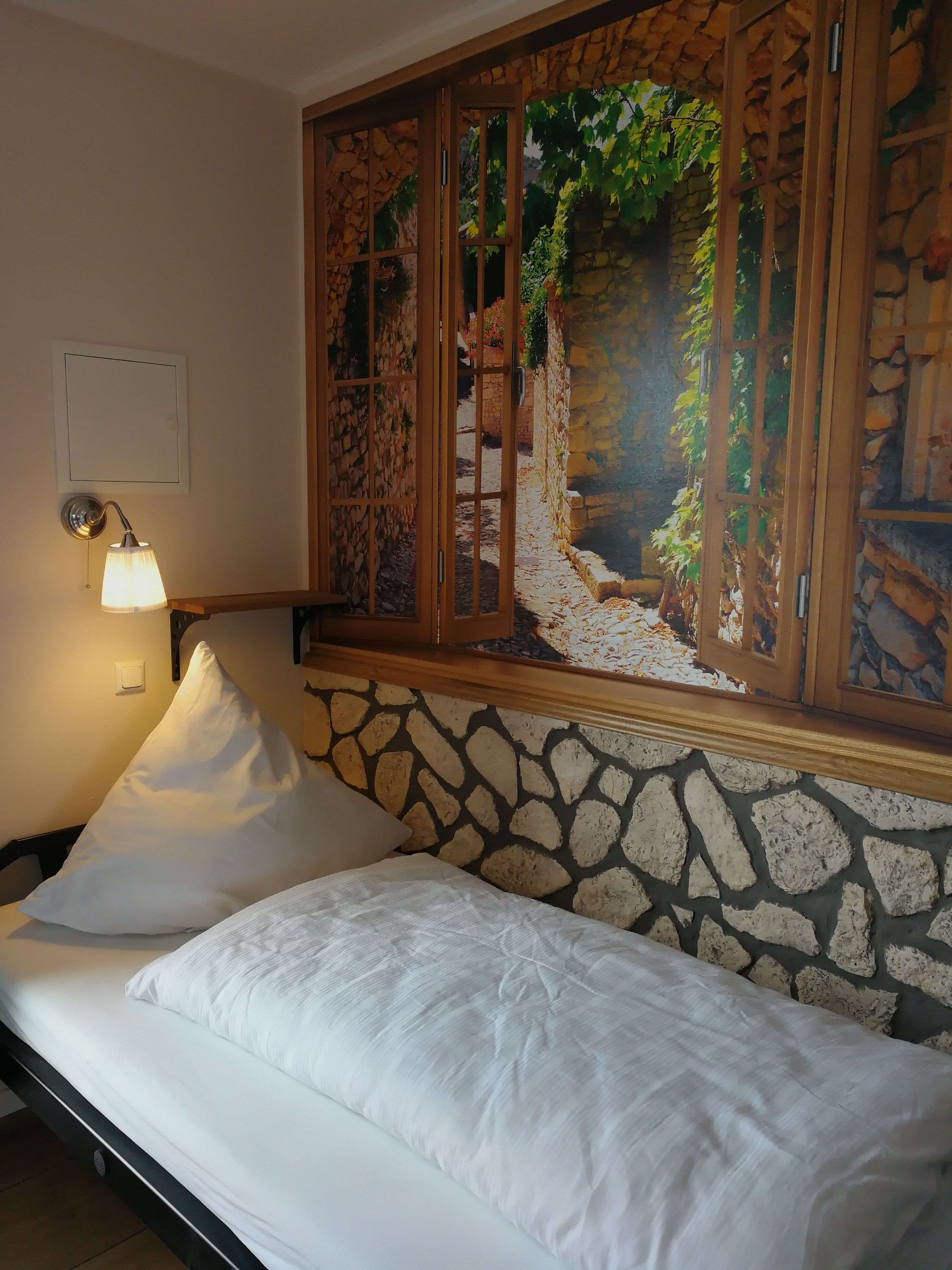 A bed with white linens, a stone wall, a wooden window frame, and a wall-mounted lamp.