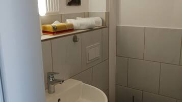 A bathroom with a white sink, toilet, and shower. There is a towel rack above the sink and a small shelf with a tissue box and a book.