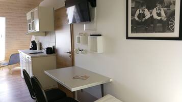 A compact kitchenette with a microwave, coffee maker, and a small sink. There is a single chair and a table with a book on it. A framed black and white photo is on the wall.