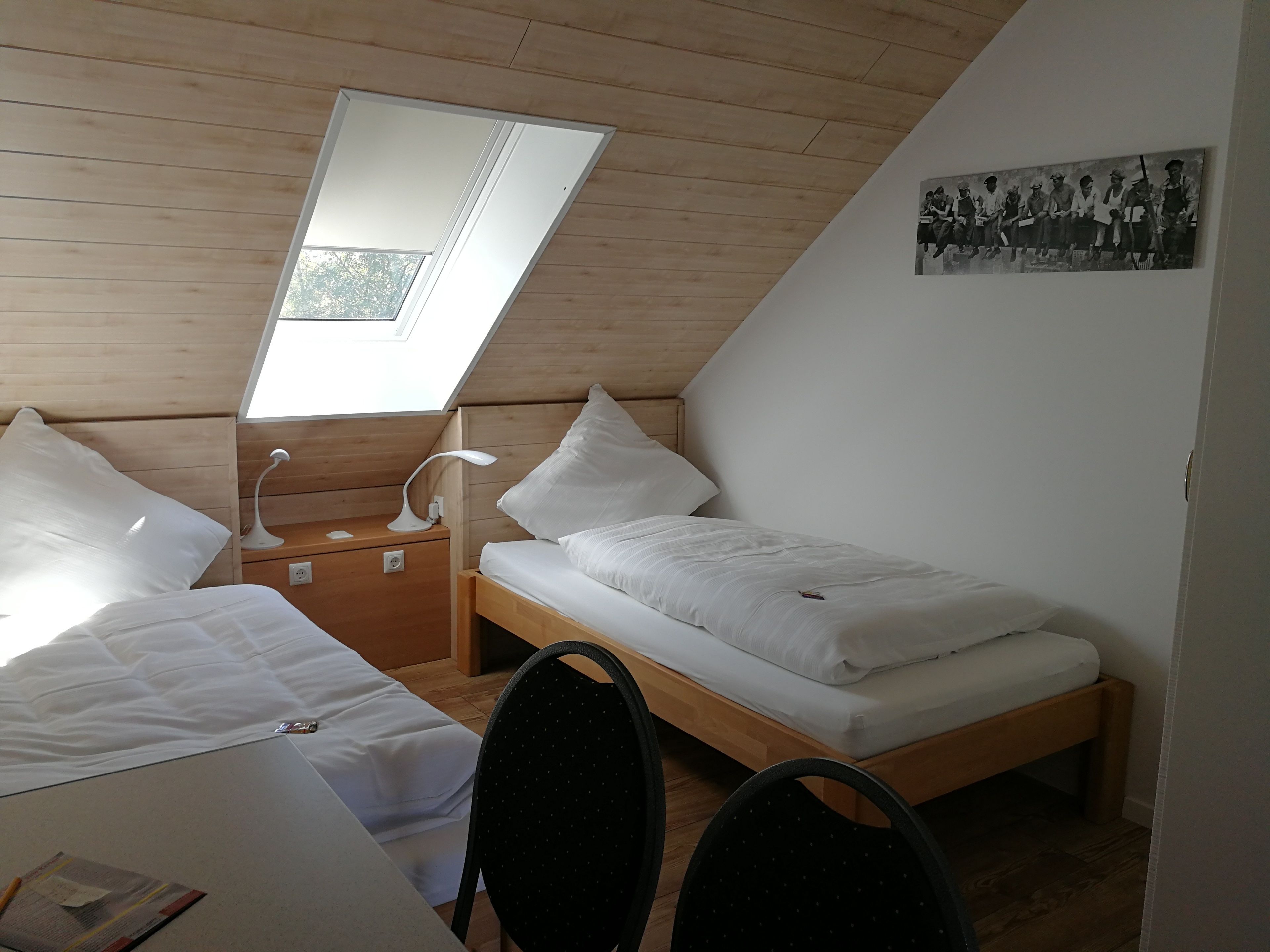 A small, well-lit bedroom with a sloped ceiling, a skylight, two single beds, a desk, and a chair.