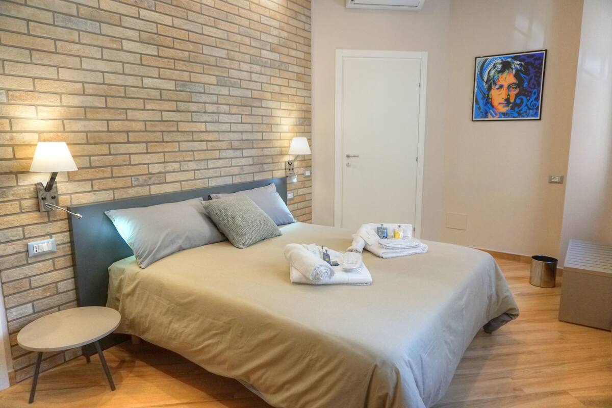A hotel room with a brick wall, a bed with a blue headboard, two bedside lamps, a small round table, and a painting on the wall.