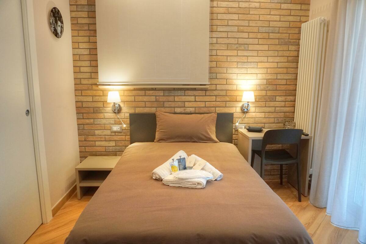 A hotel room with a brick wall, a bed with a brown bedspread, two bedside lamps, a small desk with a chair, and a radiator.