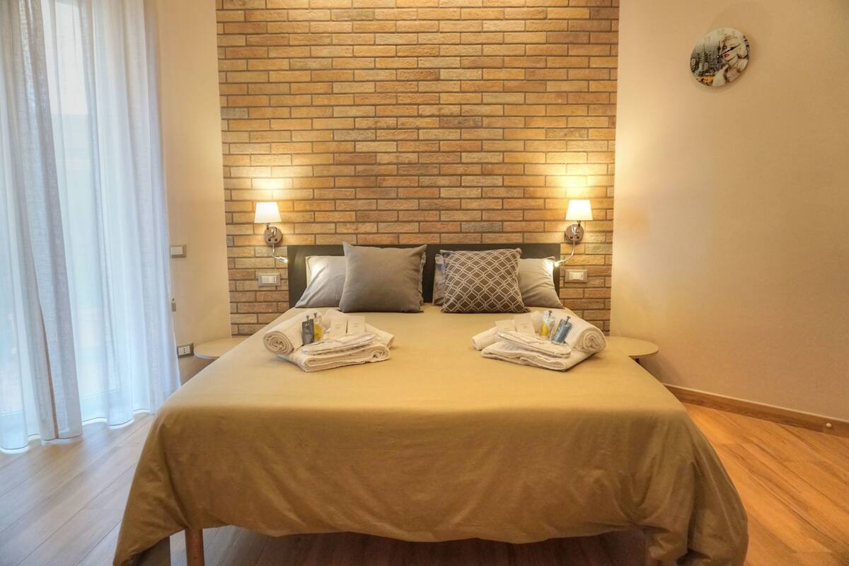 A hotel room with a brick wall, two bedside tables with lamps, a bed with a beige bedspread, and two towels on each side.