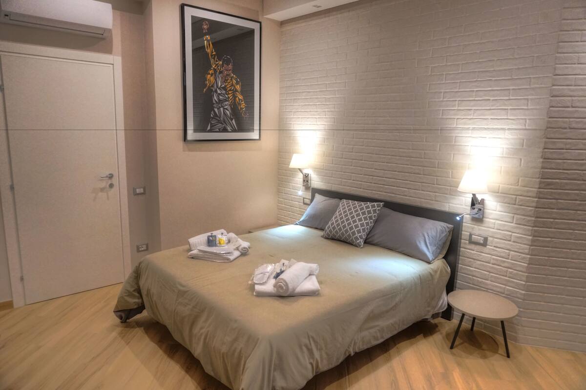 A neatly made bed with a beige blanket, a small round table, and two wall-mounted lamps.