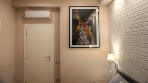 A hotel room with a door, an air conditioning unit, a framed artwork, a wall-mounted lamp, and a brick-patterned wall. - Little Rock (Gaeta)