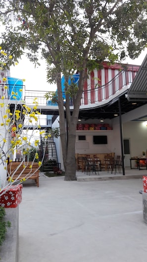 A courtyard with a tree, outdoor seating, and a building with a red and white facade. - Sweethome - Hostel (Bac Nha Trang)