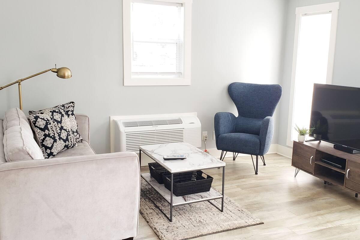 A modern living room with a sofa, armchair, coffee table, TV stand, and air conditioning unit.