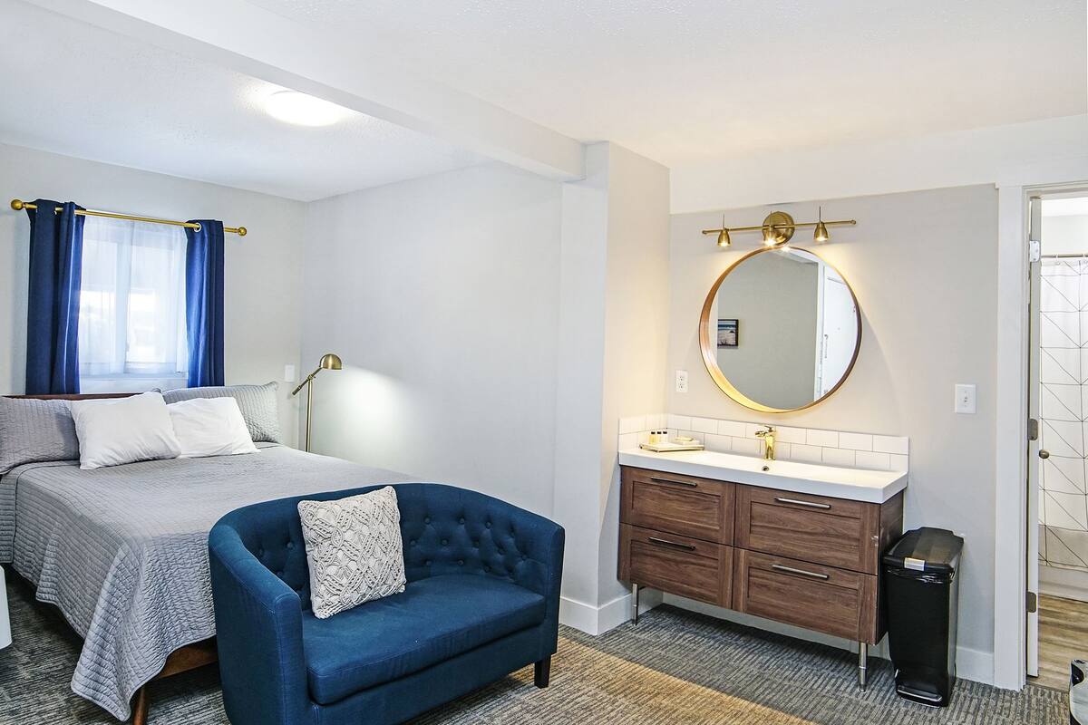 A hotel room with a bed, a blue armchair, a round mirror with lights, a white sink, and a trash bin.