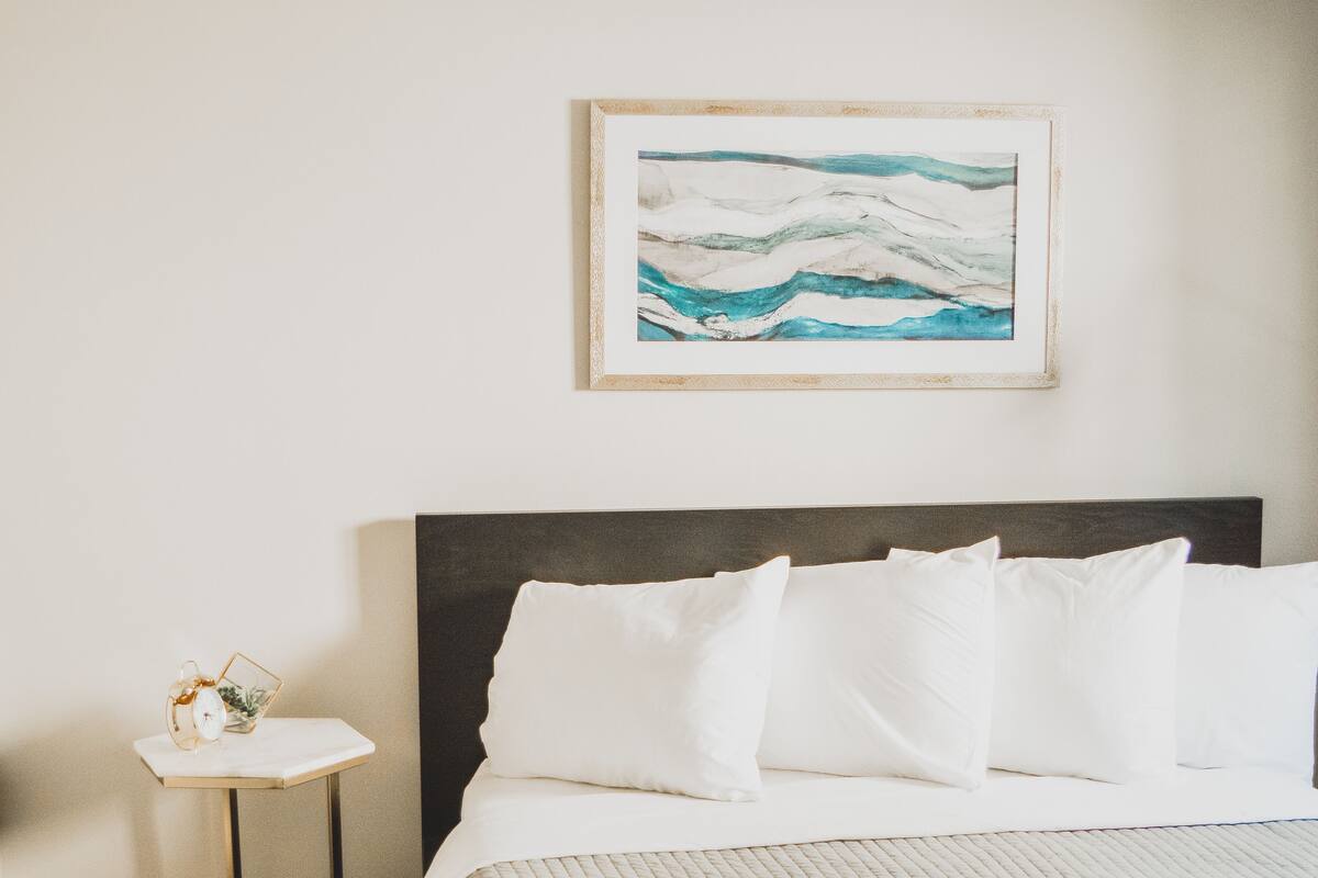 A neatly made bed with white linens and a folded towel, a small bedside table with a decorative item, and a framed artwork on the wall.