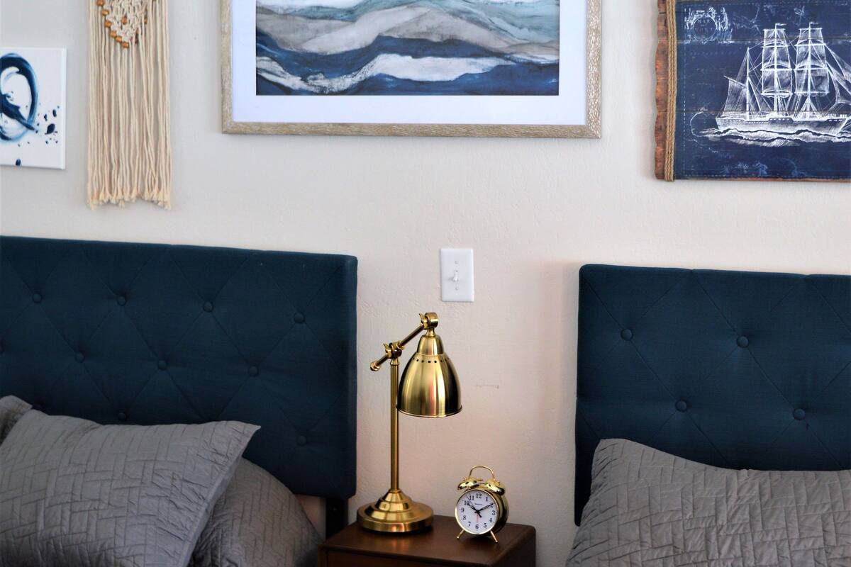 A bedroom with two beds, a wall-mounted macrame piece, framed artwork, and a small bedside table with a lamp and clock.