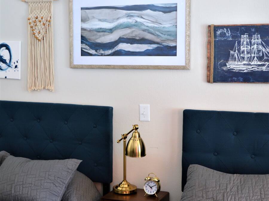 Quarto com duas camas, uma peça de macramé na parede, obras de arte emolduradas e uma pequena mesa de cabeceira com abajur e relógio.