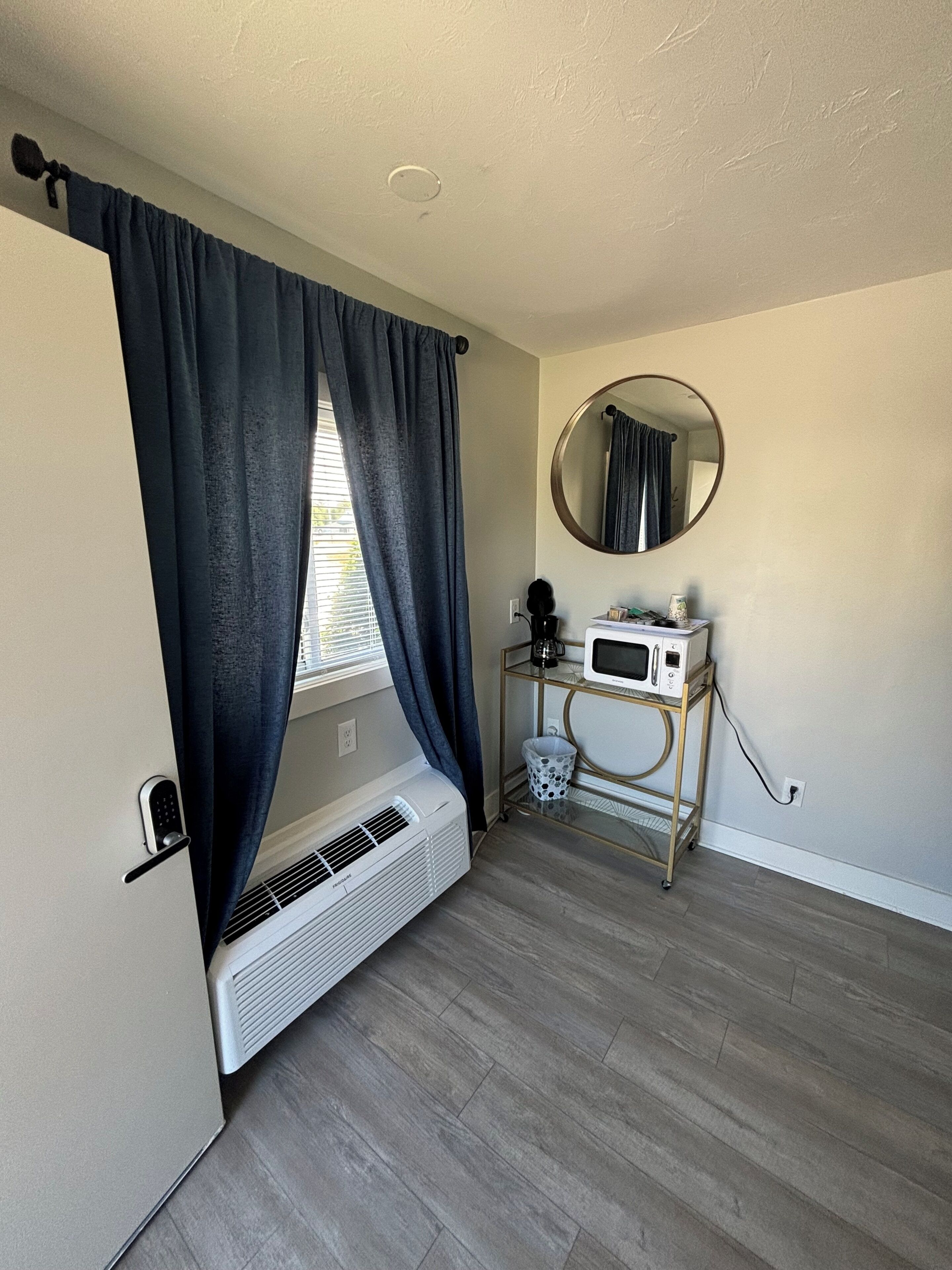 Ein Zimmer mit Fenster, einem Wandspiegel, einem weißen Heizkörper, einem kleinen Tisch mit einer Mikrowelle und einem Fußbodenlüfter.