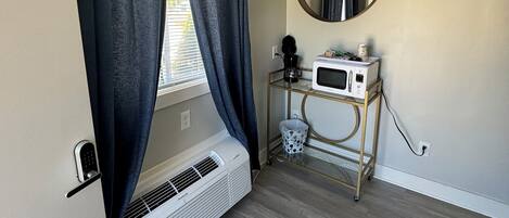 Une pièce avec une fenêtre, un miroir fixé au mur, un radiateur blanc, une petite table avec un four à micro-ondes et une grille de ventilation au sol.