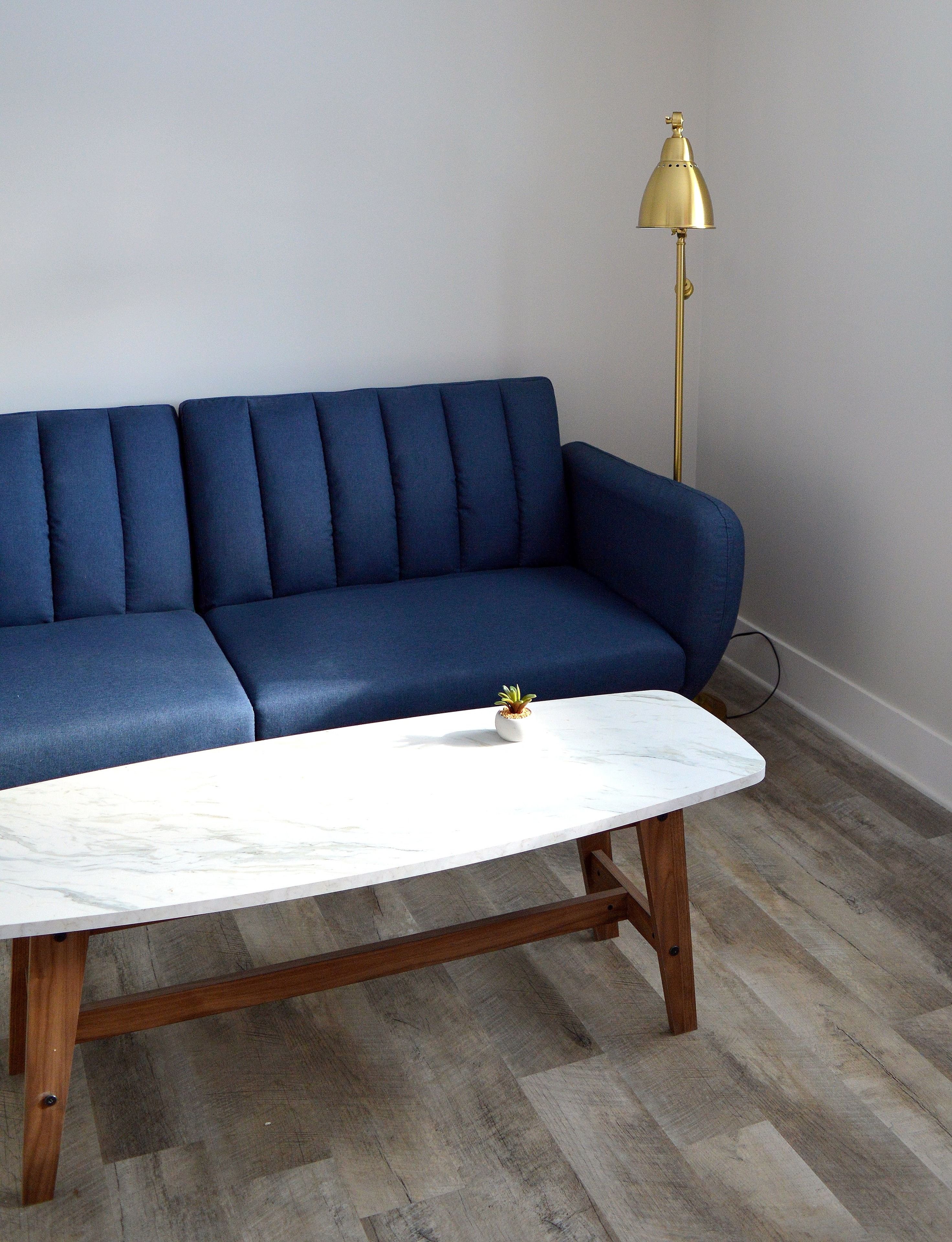 A blue sofa, a white marble coffee table, and a gold floor lamp in a room with wooden flooring.