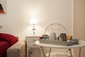 A neatly arranged hotel room with a bed, a bedside table with a lamp, a tray table with a mug, a bottle, and some snacks.