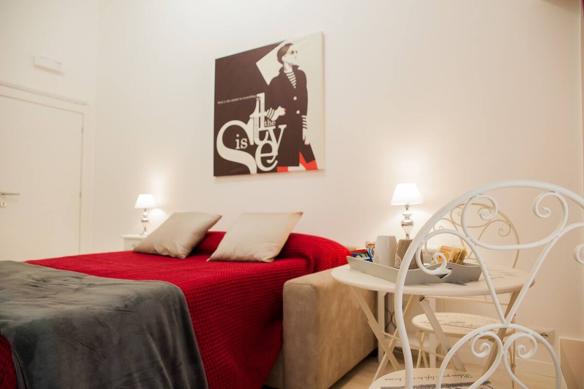 A hotel room with a red bedspread, two bedside lamps, a small white table with a tray, and a wall-mounted abstract artwork.