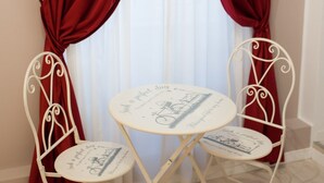 A room with two white metal chairs and a small round table, positioned in front of a window with red curtains. - Lifestyle Veneto Suite (Rome)