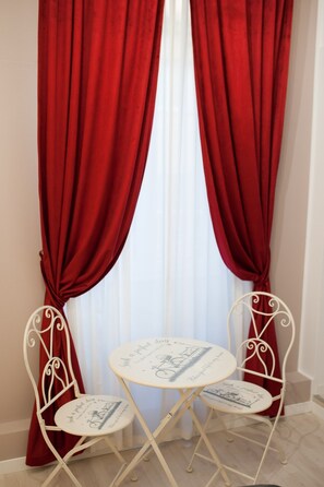 Una habitación con dos sillas blancas de metal y una pequeña mesa redonda, ubicada frente a una ventana con cortinas rojas.