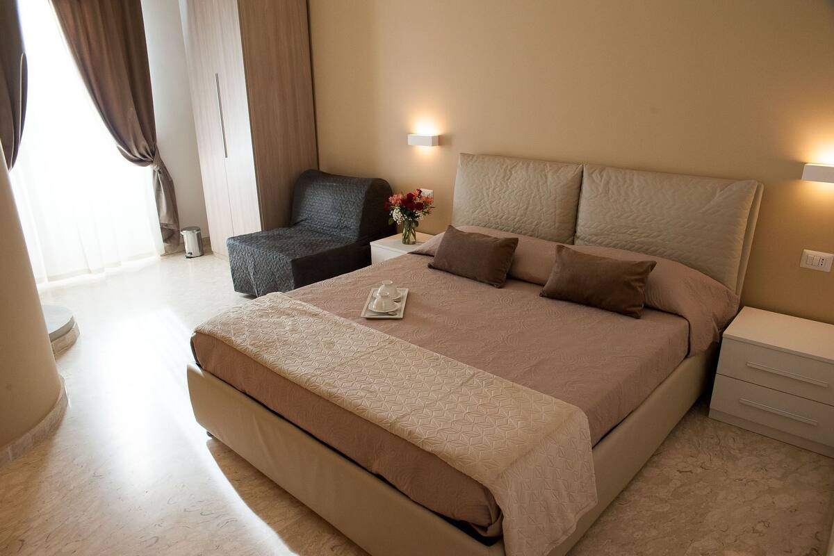 A hotel room with a large bed, bedside tables, a chair, and a small table with a vase of flowers.