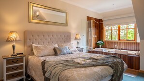 A bedroom with a large bed, two bedside tables, a mirror, a window with a view, and a bathroom with a sink and mirror. - Mountain Lodge (Mount Dandenong)