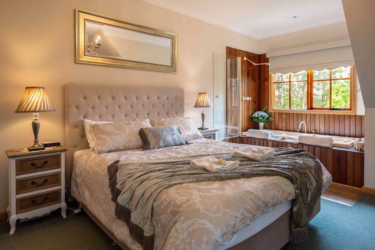 A bedroom with a large bed, two bedside tables, a mirror, a window with a view, and a bathroom with a sink and mirror.