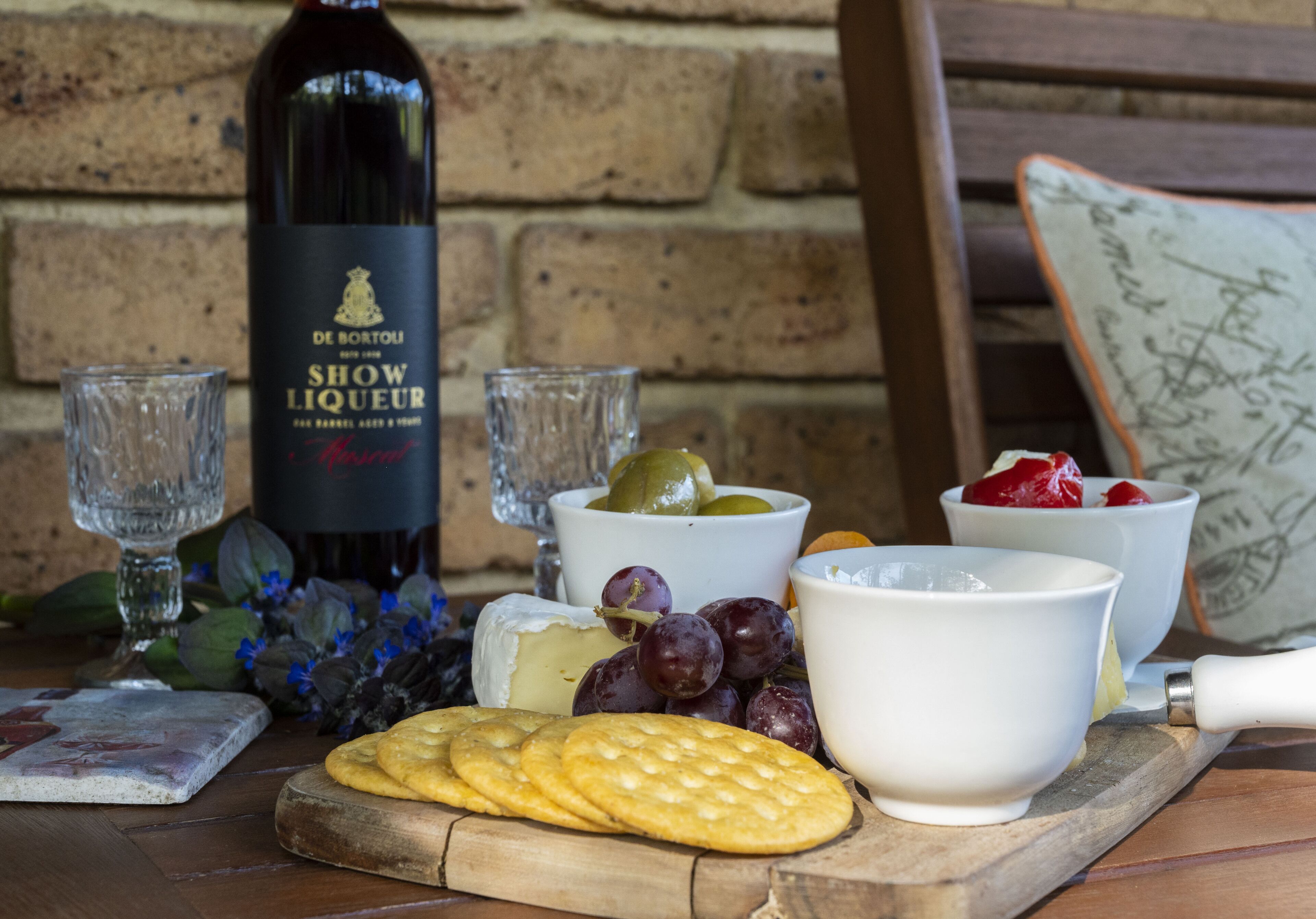 A table with a bottle of wine, glasses, cheese, grapes, crackers, and a cutting board.