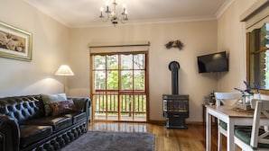 A living room with a black leather sofa, a wooden coffee table, a fireplace, a television, and a large window with a view of trees. - Mountain Lodge (Mount Dandenong)