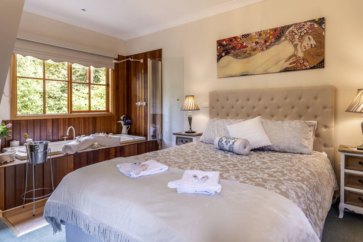 A bedroom with a large bed, a window with a view of trees, a bathroom with a sink and mirror, and a painting on the wall.