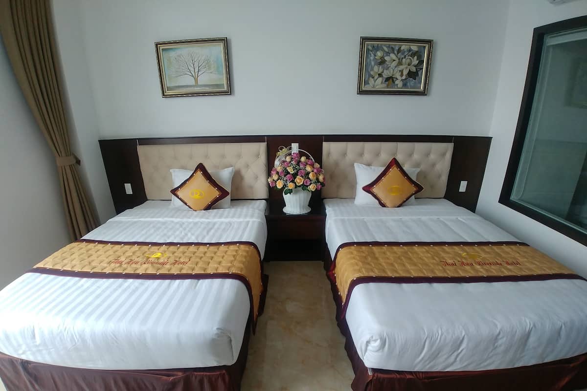 A hotel room with two beds, each with a headboard, white bedding, and decorative pillows. There are framed artworks on the walls and a vase with flowers on the nightstand.