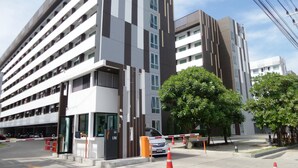 A modern multi-story building with a parking garage entrance, a gated driveway, and a tree-lined street. - PLUM Condo Phaholyothin 89 (Thanyaburi)