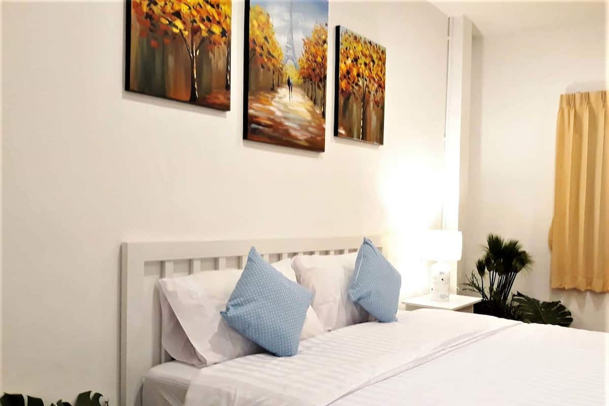 A neatly made bed with white linens and blue pillows, a white headboard, and a nightstand with a lamp and a small plant.